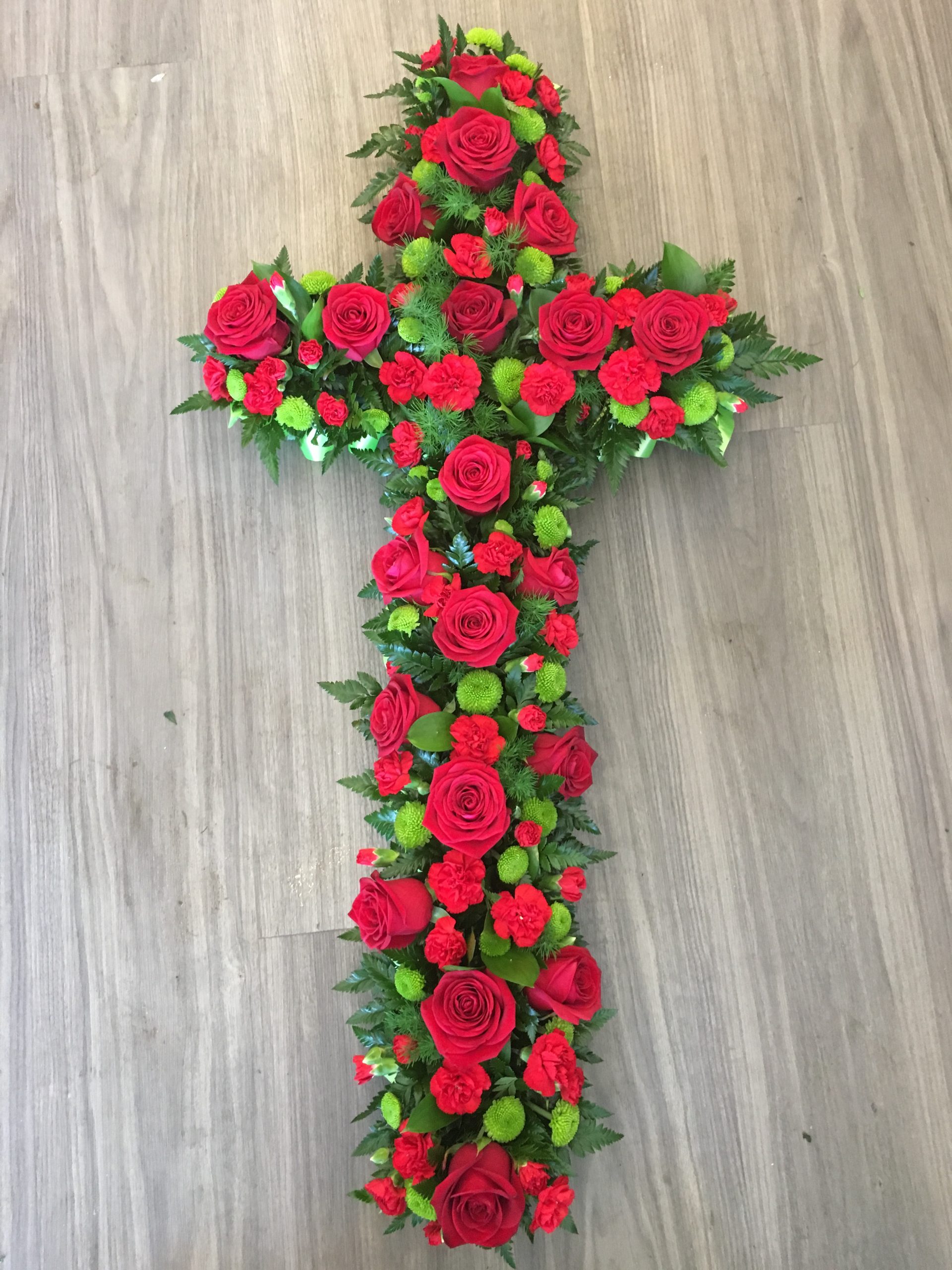 An elegant open cross tribute in red with lush foliage. Flower colour of your choice. 24 hours notice.